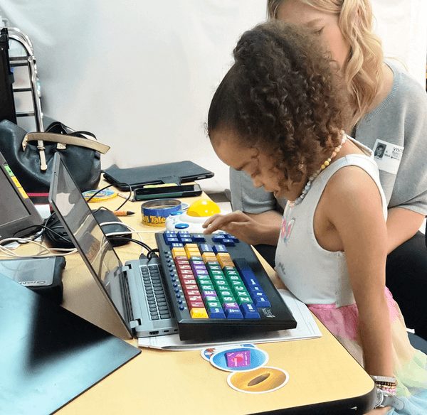 A young girl uses a colorful adaptive keyboard connected to a laptop, with an adult sitting beside her, in a classroom or therapy setting.