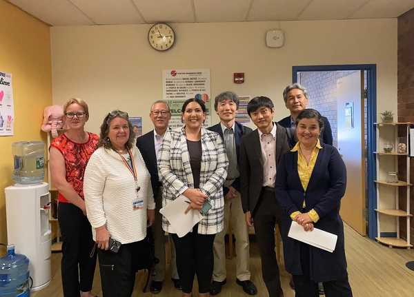 Eight people of various genders, races, and abilities standing in an office by a water cooler posing for a photo and smiling .