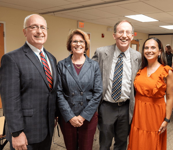 Four white people wearing formal attire stand together posing for a photo and smiling. .