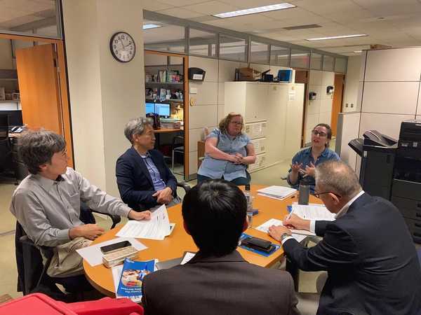 Six people of various genders, races, and abilities sitting around a table in a meeting, taking notes and talking.