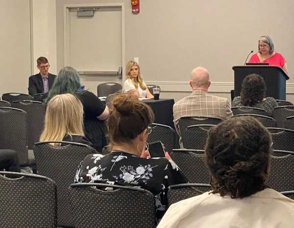 A white women with a pink shirt is presenting in front of several seated audience memners.