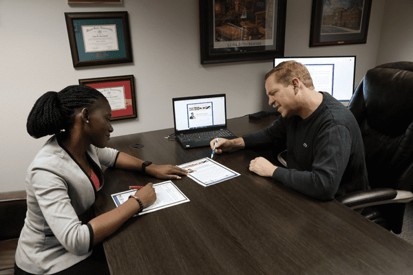 A Ugandan woman and a white man sit on either sides of a desk and review documents.